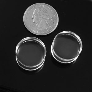 May include: Two clear acrylic round plugs, each with a slightly raised edge, are displayed on a black surface. A US quarter is included for size comparison.