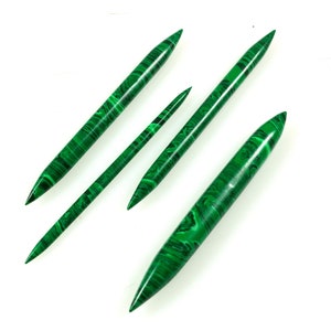 May include: Four green malachite knitting needles of varying lengths are arranged diagonally against a white background. The needles have a polished finish and display a distinctive banded pattern of dark and light green.