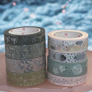 May include: Two stacks of decorative washi tape rolls. One stack features green and gray floral and bow designs. The other stack has pastel colors with clouds, stars, and moons. The bottom roll says "Sweet Dreams".