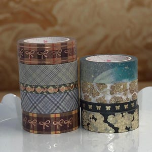 May include: Two stacks of decorative washi tape rolls. One stack features plaid, bow, and heart patterns in brown, gray, and pink. The other stack has floral, bow, and abstract designs in gold, black, and teal.