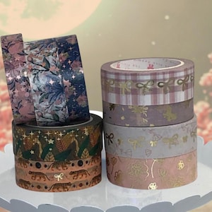 May include: A collection of decorative washi tape rolls in various designs and colors. The tapes feature floral, botanical, and geometric patterns, including tigers, bows, and plaid. The color palette includes pink, gold, green, and blue hues. The tapes are stacked on a white, scalloped-edge tray.
