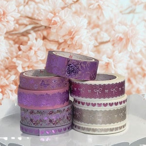 May include: A stack of decorative washi tape rolls in various shades of purple and silver. The tapes feature floral, butterfly, heart, and bow patterns. The background is a soft, blurred floral arrangement.