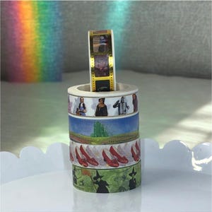 May include: A stack of decorative washi tape rolls featuring imagery from the Wizard of Oz. The rolls include images of Dorothy, the Tin Man, the Wicked Witch, and the Emerald City. One roll stands upright, displaying film strip-like images.