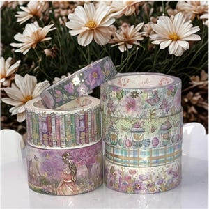 May include: A stack of decorative washi tape rolls with floral, book, and garden-themed designs. The tapes feature pastel colors, gold accents, and illustrations of flowers, butterflies, and a woman in a dress. The background includes white flowers.