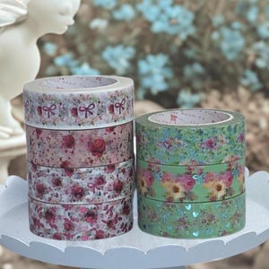 May include: A stack of decorative washi tape rolls in various floral and bow designs. The rolls feature colors like pink, green, and white. The tape is arranged on a white, scalloped-edge tray.