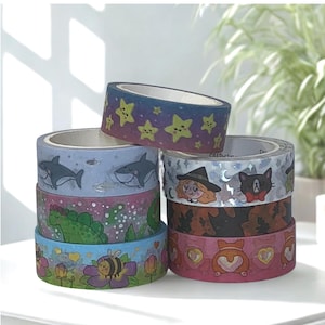 May include: A stack of decorative washi tape rolls. The rolls feature various designs, including sharks, dinosaurs, bees, stars, a witch, and a cat. The color palette includes blue, pink, yellow, and green.