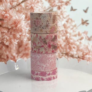 May include: Four rolls of decorative washi tape stacked on a white tray. The tape features floral and butterfly patterns in shades of pink, beige, and brown. The background includes pink flowers and butterfly cutouts.