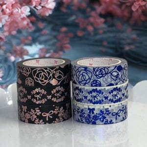 May include: Two stacks of decorative washi tape rolls. One stack features black tape with rose gold floral and bow designs. The other stack has blue and white tape with floral and bow patterns. The text "The Pink Room Co." is visible on the tape.