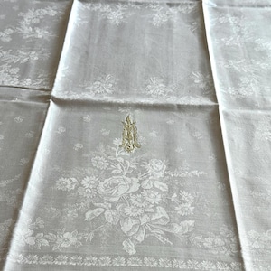 May include: An off-white tablecloth, folded and displaying a floral pattern and a central embroidered monogram. The design includes roses and other flowers, with a decorative border. The fabric has a subtle sheen.