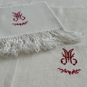 May include: Two white linen napkins with red embroidered initials "M" and a small floral design. The napkins have a white fringe border.