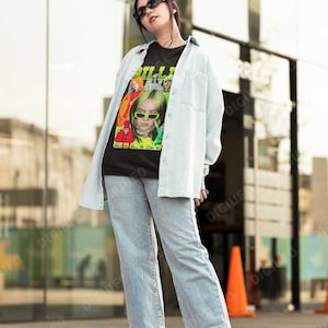 May include: A person wearing a black t-shirt with a green and white graphic of Billie Eilish. The t-shirt has the text "Billie Eilish" in green letters. The person is also wearing light blue jeans and white sneakers. They are standing in front of a building with large windows.
