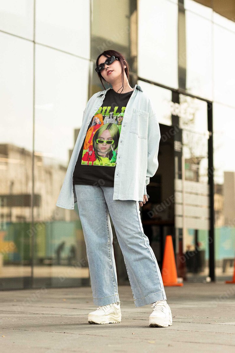 May include: A person wearing a black t-shirt with a green and white graphic of Billie Eilish. The t-shirt has the text "Billie Eilish" in green letters. The person is also wearing light blue jeans and white sneakers. They are standing in front of a building with large windows.