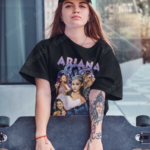 May include: A black t-shirt with a graphic print of Ariana Grande. The graphic features multiple images of Ariana Grande in different poses and outfits. The text "Ariana" is printed in purple letters above the graphic.