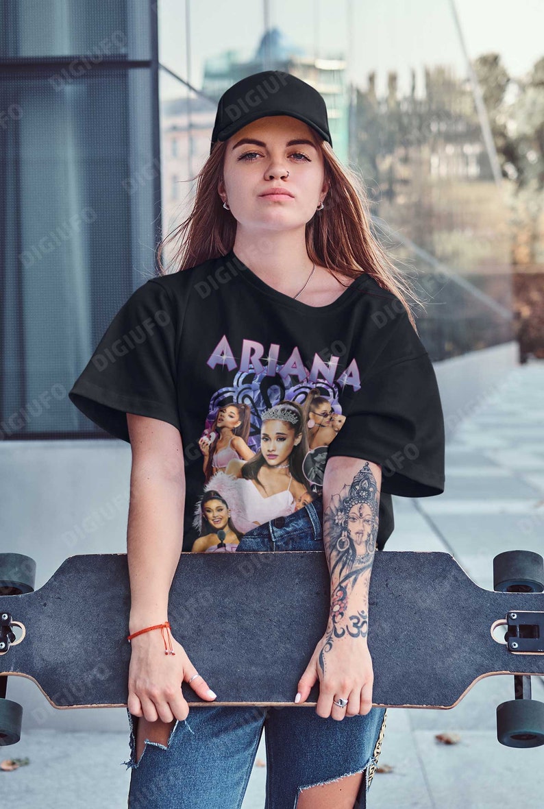 May include: A black t-shirt with a graphic print of Ariana Grande. The graphic features multiple images of Ariana Grande in different poses and outfits. The text "Ariana" is printed in purple letters above the graphic.