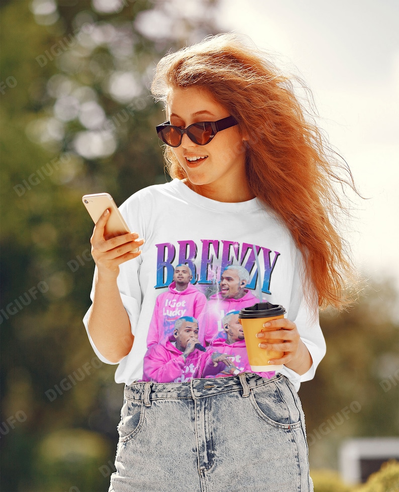 May include: White t-shirt with a graphic of a singer in a pink hoodie, the word "Breezy" in blue and purple, and the text "I Got Lucky". The shirt is worn by a person wearing sunglasses and jeans.
