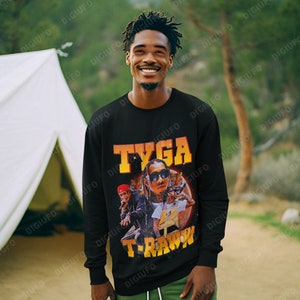 May include: Black long-sleeve sweatshirt featuring a graphic of Tyga with the text "TYGA" and "T-RAWW" in orange and yellow. The person is wearing green cargo pants and is smiling in an outdoor setting.