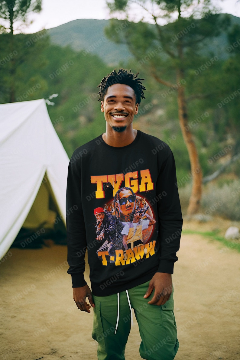 May include: Black long-sleeve sweatshirt featuring a graphic of Tyga with the text "TYGA" and "T-RAWW" in orange and yellow. The person is wearing green cargo pants and is smiling in an outdoor setting.