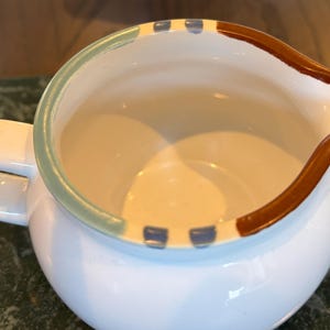 以下が含まれることがあります： 茶色、青、緑の縁取りが施された白いセラミック製のクリーマー。クリーマーは丸みを帯びたボディとハンドルが付いています。
