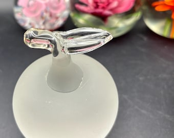 May include: A frosted glass bottle stopper with a clear, wing-shaped handle. The stopper sits in front of three glass paperweights, each containing a different flower in shades of pink and orange.