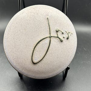 Könnte beinhalten: Ein runder, hellgrauer Keramikdeckel mit einem dunkelgrünen, abstrakten Design. Der Deckel hat einen leicht erhöhten Rand und wird auf einem schwarzen Metallständer präsentiert. Das Design scheint handgemalt zu sein.