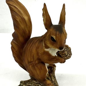 May include: A detailed ceramic squirrel figurine, primarily brown with white accents, holding a walnut. The squirrel is perched on a tree branch, showcasing a realistic design. The figurine is a decorative item.