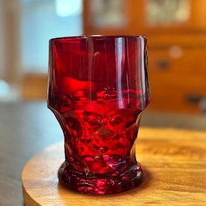 May include: A vibrant red glass tumbler with a textured design. The glass is set on a wooden coaster with the word "CARAVAN" engraved on it. The tumbler's color is a deep, rich red, and it appears to be made of thick glass.