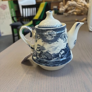 May include: A cream-colored ceramic teapot featuring a blue transferware pattern depicting a rural landscape with cottages, trees, and birds. The teapot includes a matching lid with a small knob, a curved handle, and a spout, all decorated with blue details.