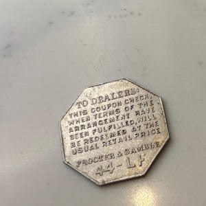 Puede incluir: Un cheque de cupón plateado para Procter & Gamble. El cupón tiene forma octogonal y tiene el texto "TO DEALERS: THIS COUPON CHECK, WHEN TERMS OF THE ARRANGEMENT HAVE BEEN FULFILLED, WILL BE REDEEMED AT THE USUAL RETAIL PRICE PROCTER & GAMBLE 44-LF" impreso en él.