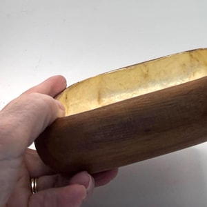 May include: A handcrafted wooden bowl with a smooth, dark brown exterior and a light yellow interior. The bowl has an elongated, oval shape and is held by a hand. This decorative bowl is suitable for serving or display.