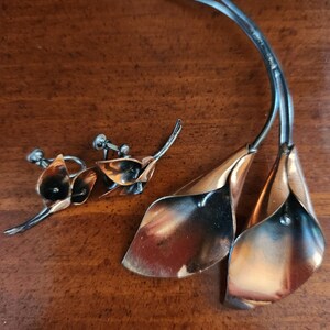 May include: A set of copper and silver tone jewelry featuring a brooch and earrings. The brooch is shaped like three stylized calla lilies. The earrings are smaller versions of the lilies.
