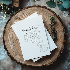 Può includere: Un libro bianco con il testo "baby's first woodland ABC book" e un'illustrazione in bianco e nero di un orso che tiene un fiore.