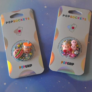 Puede incluir: Dos agarres para teléfono PopSockets PopGrip con tapas intercambiables. El primer agarre presenta un personaje de dibujos animados rosa y blanco con un cono de helado rosa y blanco. El segundo agarre presenta un hot dog, un té de boba y un personaje de dibujos animados verde. Ambos agarres tienen un fondo azul con purpurina blanca y rosa.