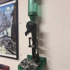 May include: A reloading press mounted on a wall. The press is primarily black and green, with a clear, cylindrical powder reservoir. The base is green, and the press has various mechanical components. A framed picture hangs on the wall to the left.