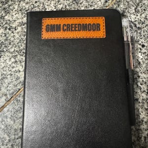 May include: Black leather notebook with a brown leather patch that says "6MM CREEDMOOR" on the front. A black pen is attached to the notebook.