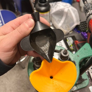 Könnte beinhalten: Ein schwarzer Plastiktrichter mit einem kleinen Loch am Boden wird verwendet, um kleine, silberfarbene Pellets in einen gelben Plastiktrichter zu füllen. Der gelbe Trichter ist an einer grünen Ladepresse befestigt.