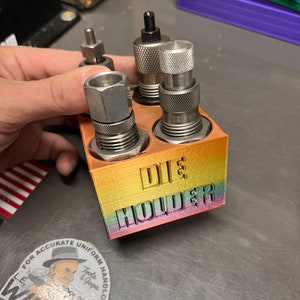 May include: A rainbow-colored die holder with the word "DIE HOLDER" printed on the front. The holder has four slots for dies, and there are four dies in the slots. The dies are made of metal and have a silver finish.