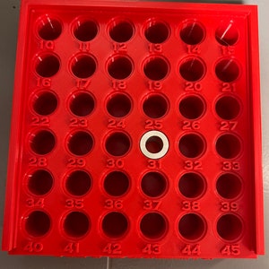 May include: A red plastic tray with 45 circular holes, each numbered. A white ring sits in hole number 31. The tray is designed to hold small objects, likely for organization or storage.