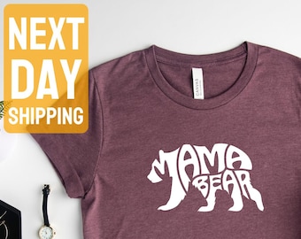 May include: A heather maroon t-shirt with a white graphic of a bear silhouette with the words "Mama Bear" inside the bear. The shirt is laid flat on a white surface.