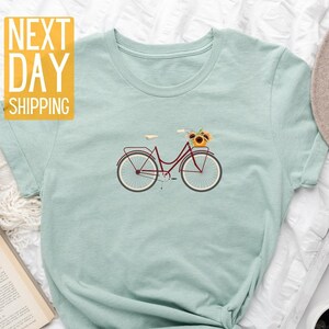 May include: A light blue t-shirt with a graphic of a bicycle with a basket of sunflowers. The text "NEXT DAY SHIPPING" is in yellow on a white background.