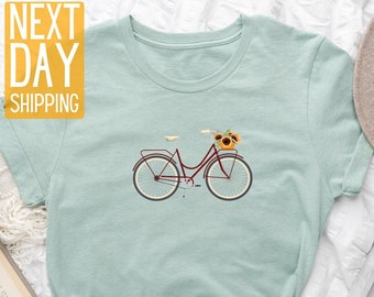 May include: A light blue t-shirt featuring a graphic of a bicycle with a basket of sunflowers. The text "NEXT DAY SHIPPING" is in yellow on a white background.