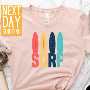 May include: A pink t-shirt with a graphic of colorful surfboards and the word "SURF" in a retro style.