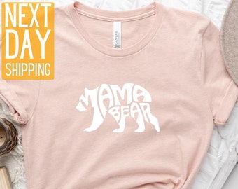 May include: A pink t-shirt featuring a white bear silhouette design. The bear is formed from the words "Mama Bear".
