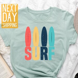 May include: A light blue t-shirt with a retro-style graphic of four colorful surfboards and the word "SURF" in bold letters.