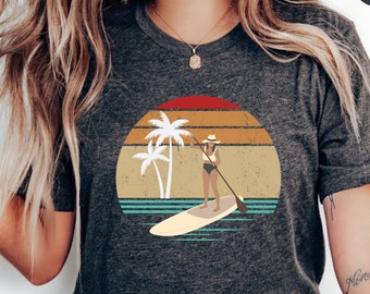 May include: A heather dark grey t-shirt with a vintage-style graphic of a person paddleboarding in front of palm trees and a sunset. The text "Mon Océan" is tattooed on the person's arm.
