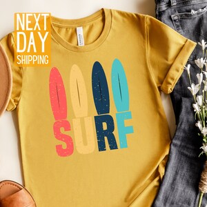 May include: A mustard yellow t-shirt with a retro-style graphic of three surfboards and the word "SURF" in colorful block letters.