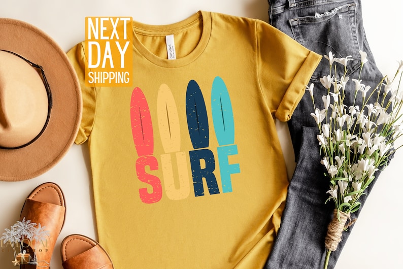 May include: A mustard yellow t-shirt with a retro-style graphic of three surfboards and the word "SURF" in colorful block letters.