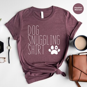 May include: Heather maroon t-shirt with the text "DOG SNUGGLING SHIRT" and a white paw print with a small heart in the center.