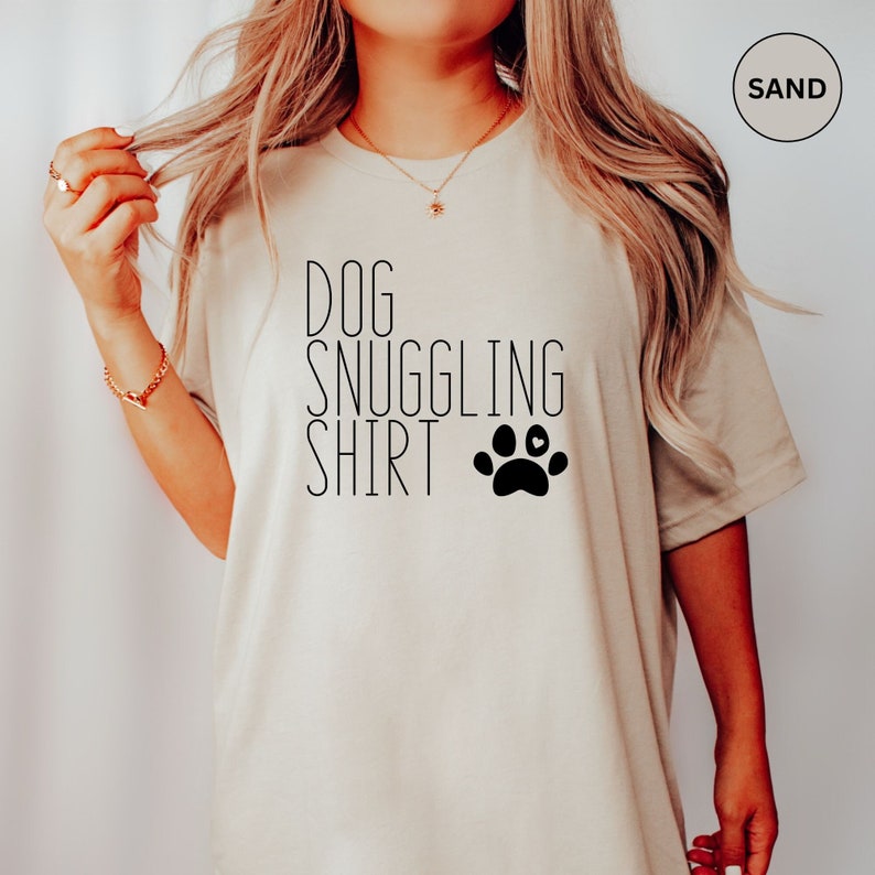May include: A beige t-shirt with the text "DOG SNUGGLING SHIRT" and a black paw print with a small heart in the center.
