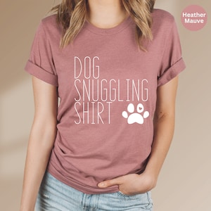 May include: A heather mauve t-shirt with the text "DOG SNUGGLING SHIRT" and a white paw print with a small heart in the center.