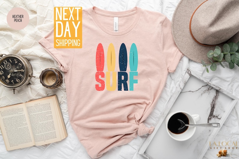 May include: A pink t-shirt with a graphic of colorful surfboards and the word "SURF" in a retro style.